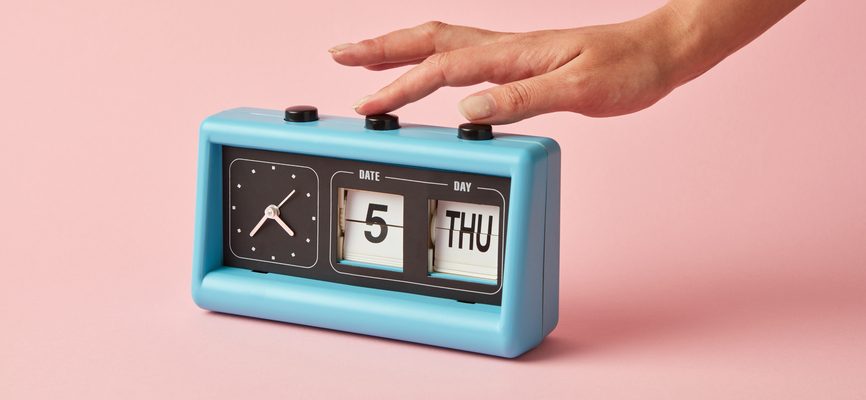 A blue flip oclock shows date 5, day Thuesday, time twenty two to five on a color of the year 2019 Living Coral pantone background. Retro style. Womans hand pushes on a buttom.